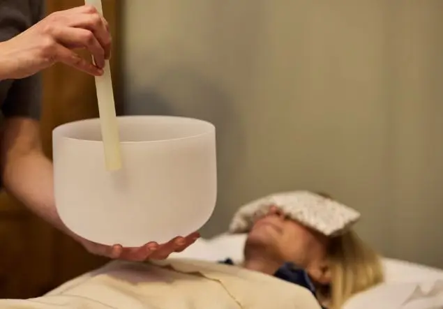 A woman with covered eyes lies on a bed while hands in the foreground are using a singing bowl for Reflexology