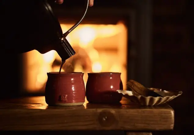 A teapot pouring liquid into two mugs by a roaring fire