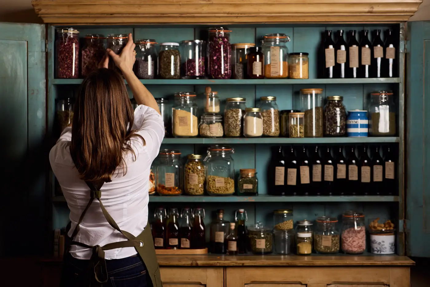 A woman (Kate Goodwin, The Soul Polisher) organises herbs in a sideboard full of glass jars