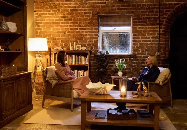 A woman in a pink dress is explaining something from a document in her hand to another woman. They are in a warm room with candles and wooden furniture