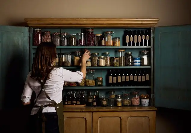 A woman reaches for a glass jar of herbs from a sideboard full of jars